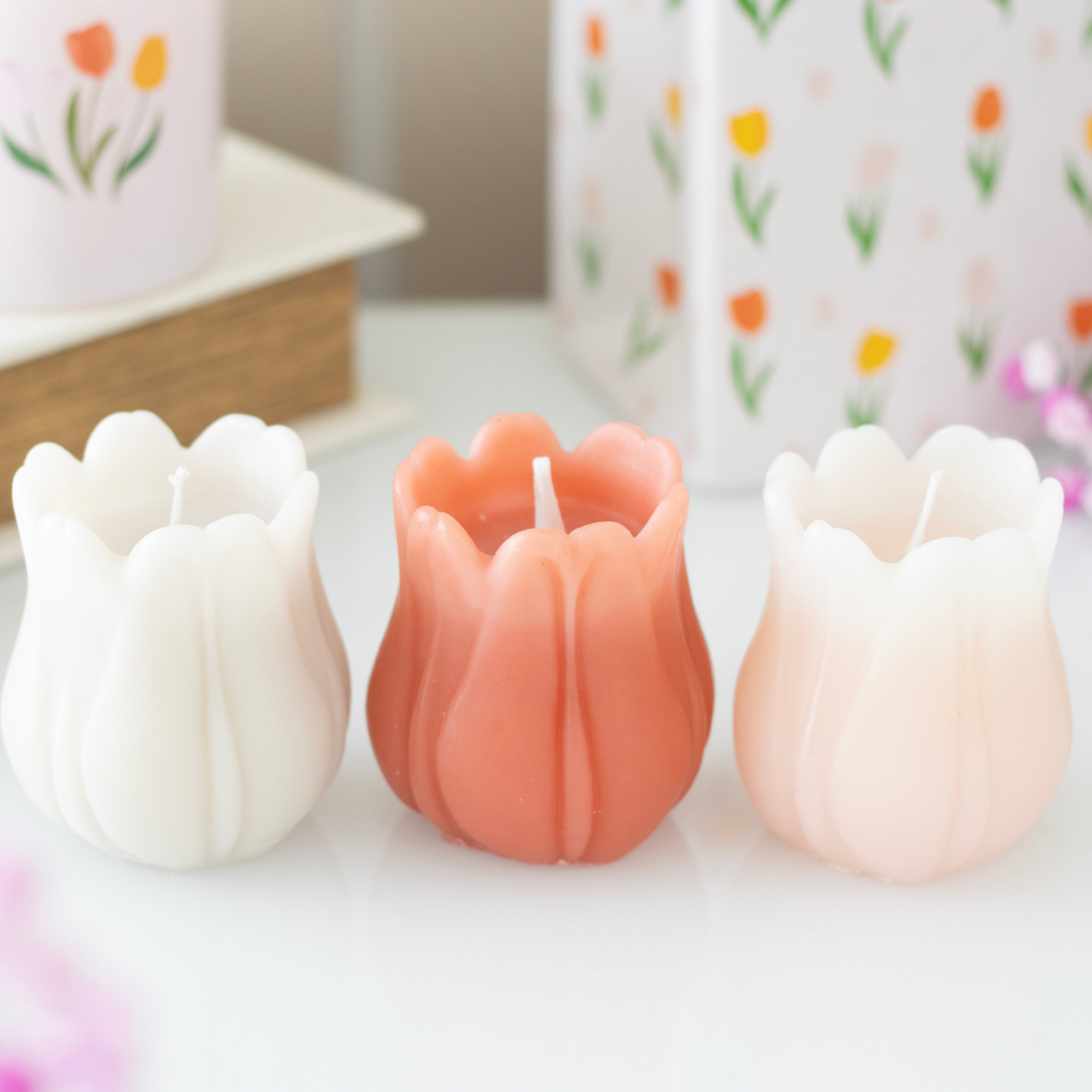 Set of 3 tulip-shaped candles in white, coral, and peach tones, displayed on a white surface with floral packaging in the background.
