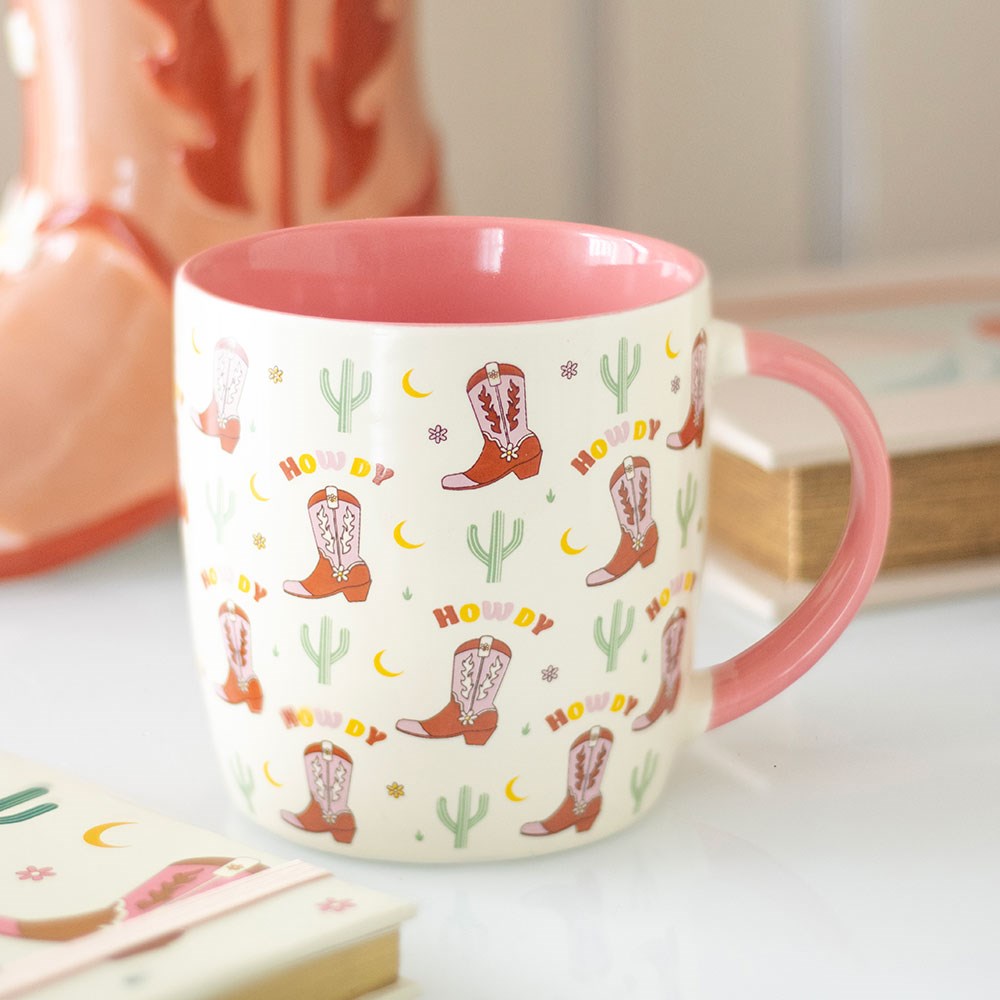Cowboy Print Coffee Mug featuring boots, cactus, and “Howdy” design on cream ceramic, pink interior, western-style cup in gift box.
